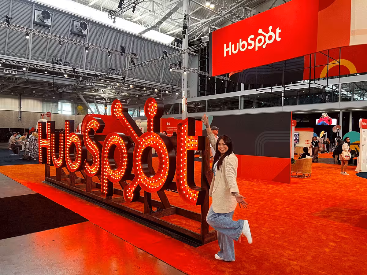 Opensense team member posing cheerfully in front of a large illuminated HubSpot sign at the INBOUND convention hall, with bright orange flooring and HubSpot branding displayed throughout the event space.
